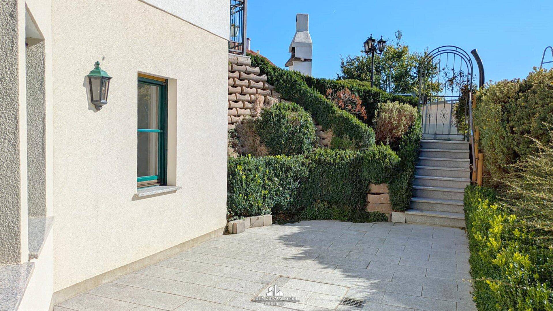 Gepflasterter Außenbereich mit Treppe, Hecken und einem Grill im Hintergrund.