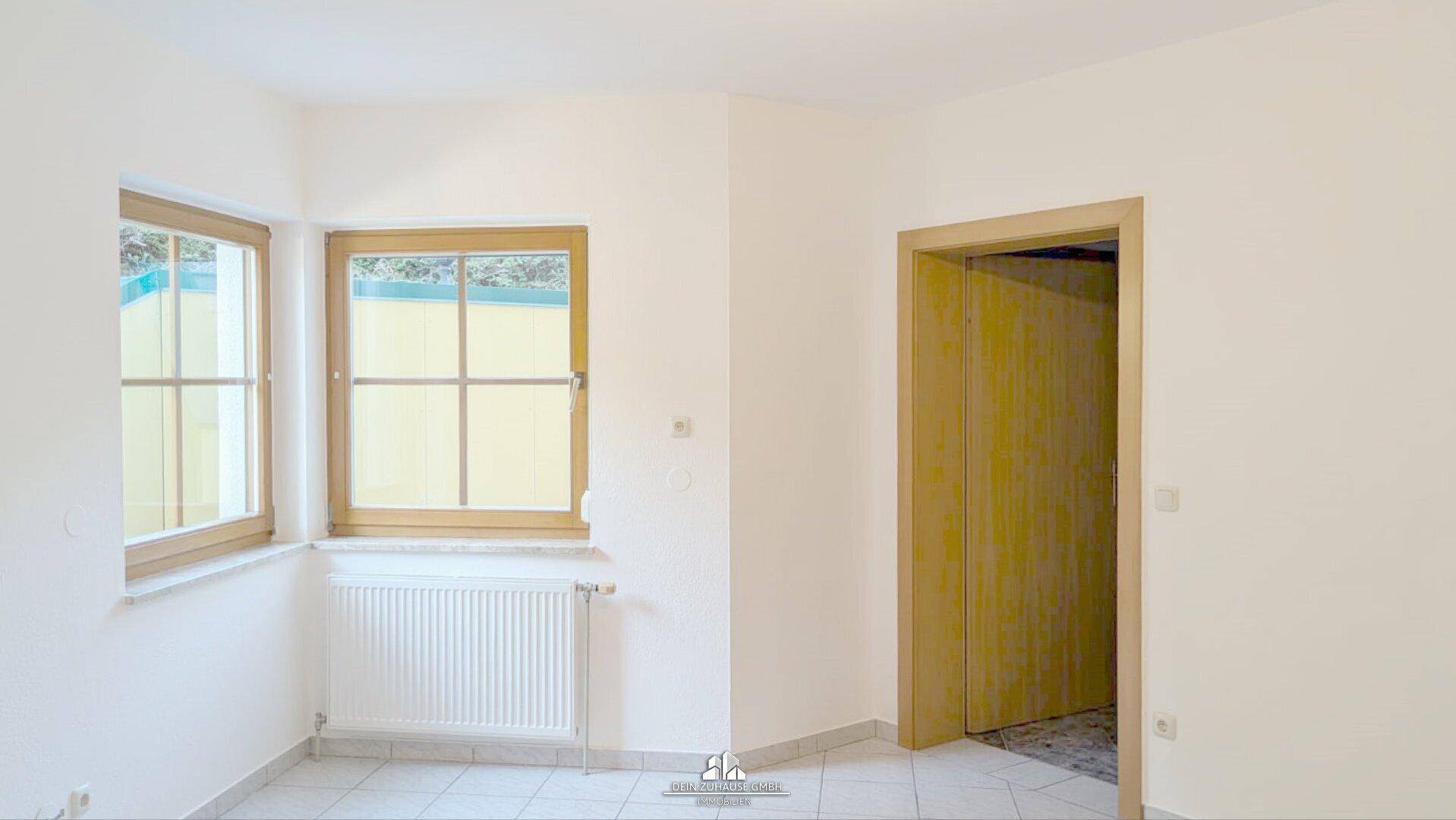 Heller Raum mit Fliesenboden, weißen Wänden und zwei Fenstern mit Holzrahmen.