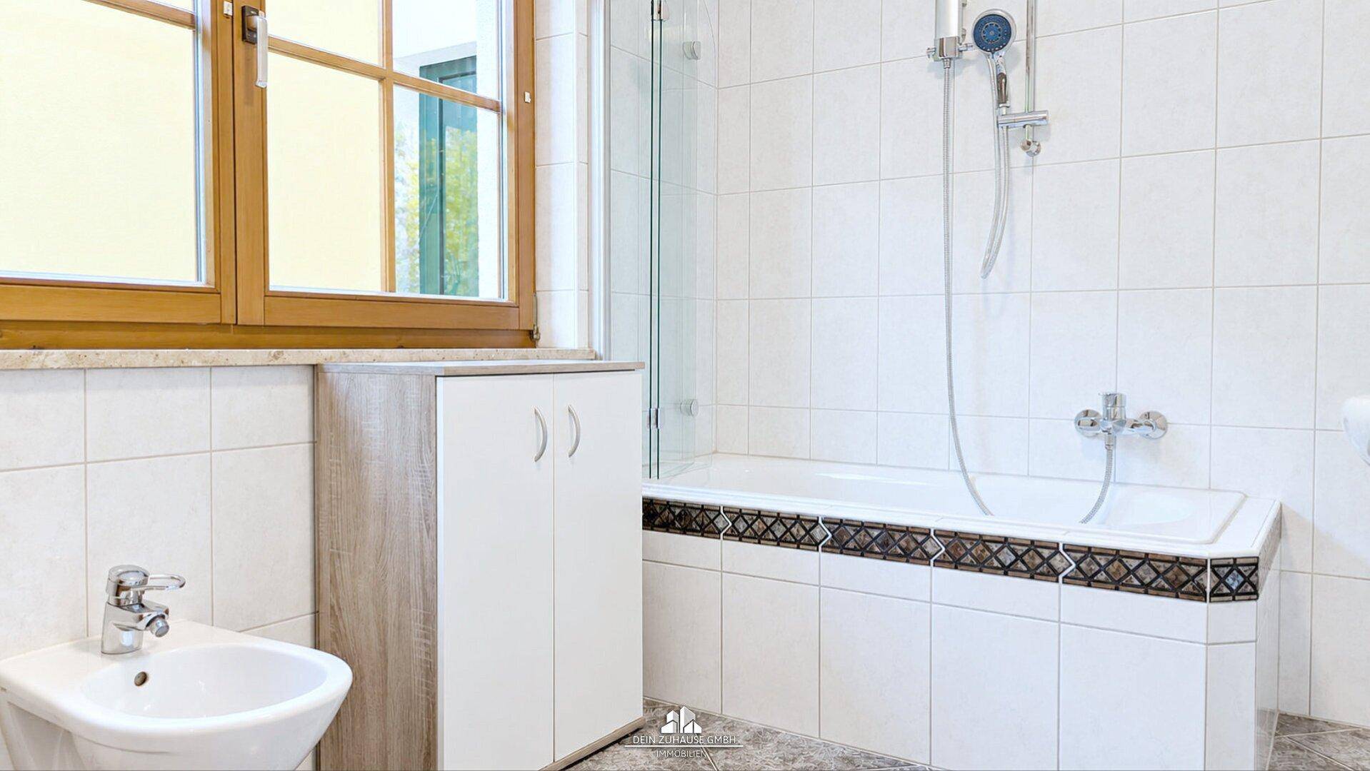 Badezimmer mit Badewanne, Bidet, Fenster und weißen Fliesen mit Dekorstreifen.
