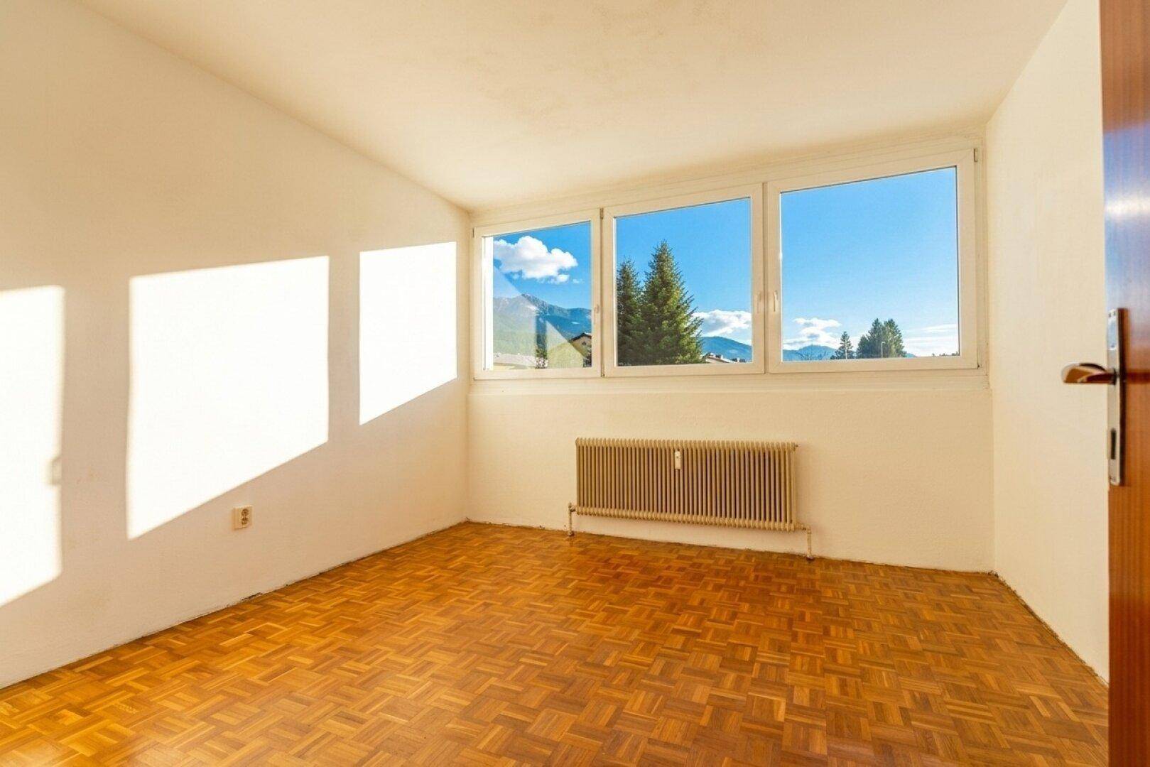 Leeres Zimmer mit Parkettboden, Heizkörper und großzügigen Fenstern mit Bergblick.