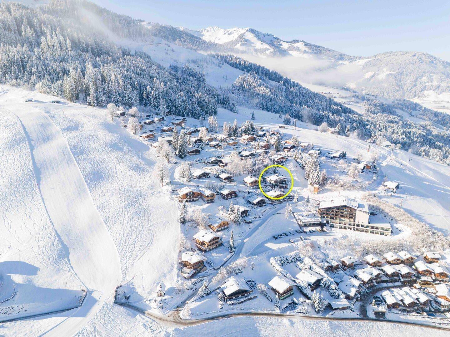 Winterliche Luftaufnahme eines Bergdorfes mit verschneiten Häusern und Skipisten.