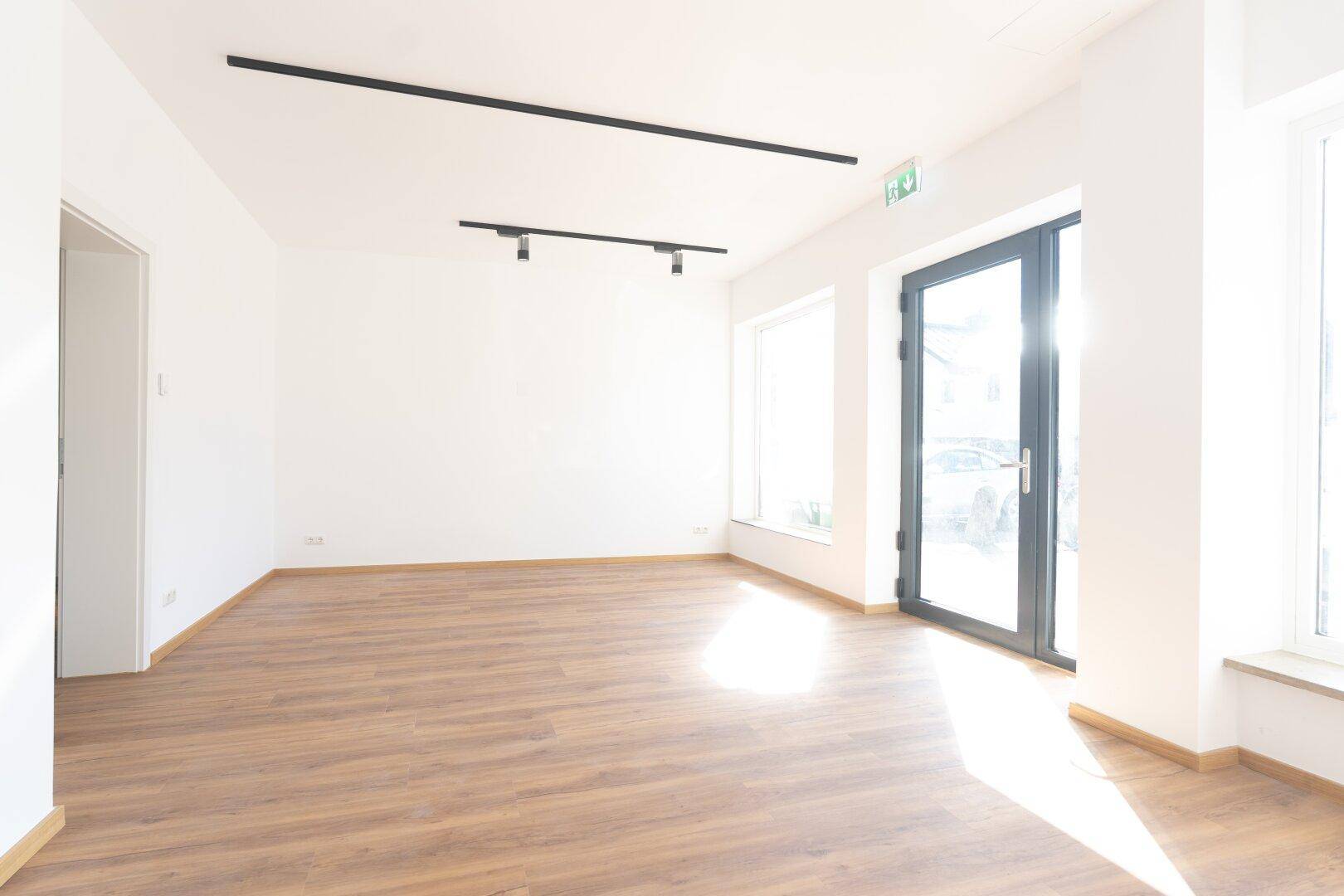 Geräumiger, heller Innenraum mit Laminatboden, weißen Wänden und einer großen Glastür zum Außenbereich.