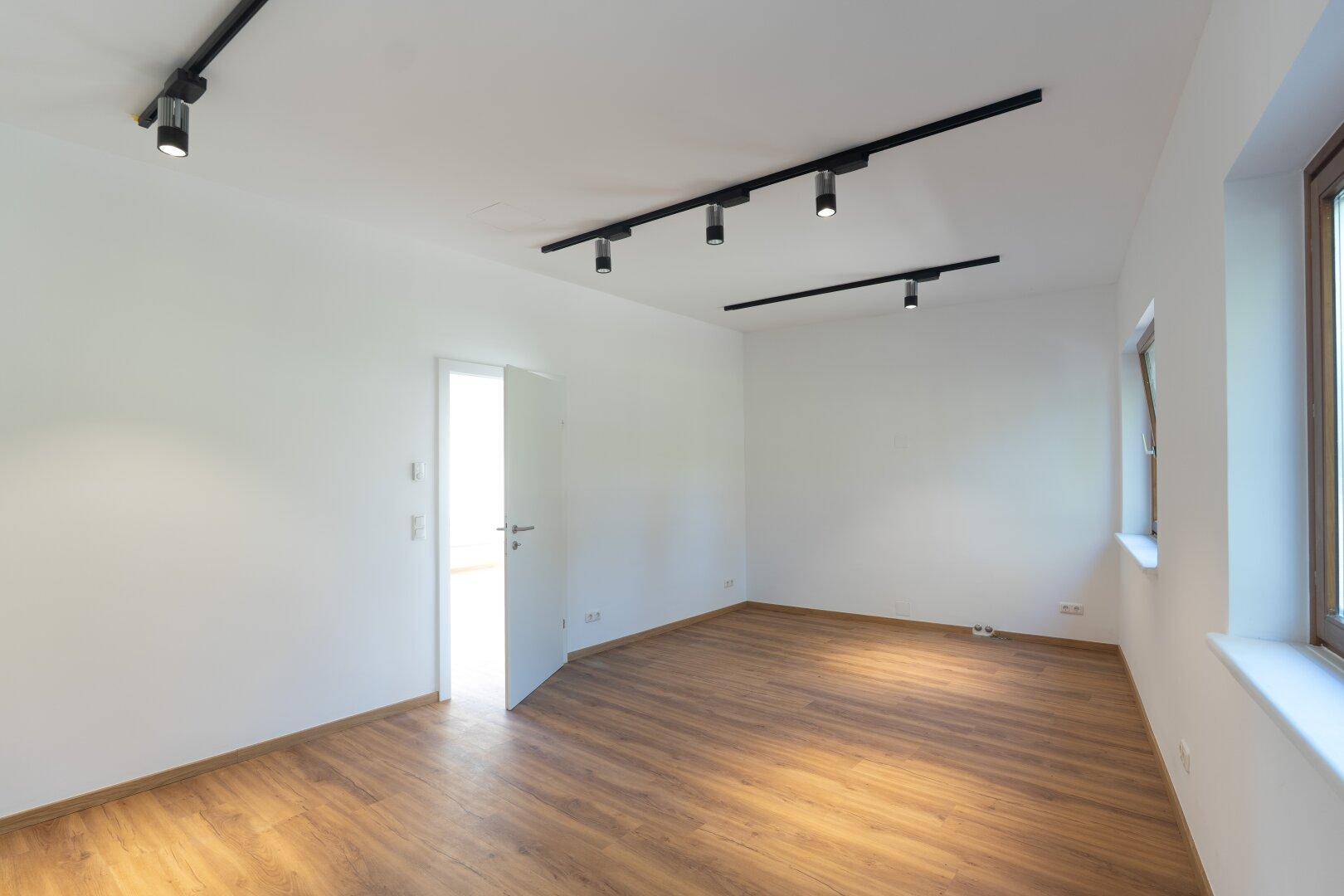 Leerer, heller Raum mit Laminatboden, weißen Wänden und einer offenen Tür, die Licht hereinlässt.