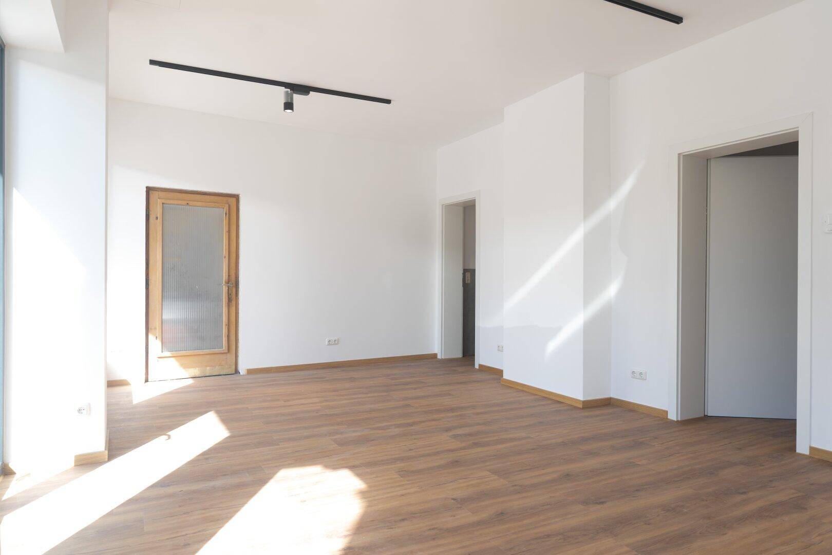 Heller, leerer Raum mit Laminatboden, weißen Wänden und mehreren Türen, die zu anderen Bereichen führen.