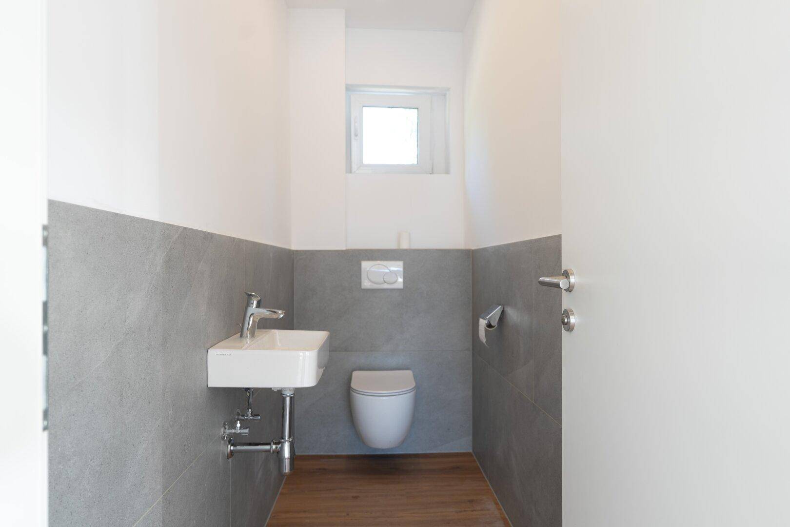 Modernes Gäste-WC mit grauen Fliesen, weißem Waschbecken, wandhängendem WC und kleinem Fenster.
