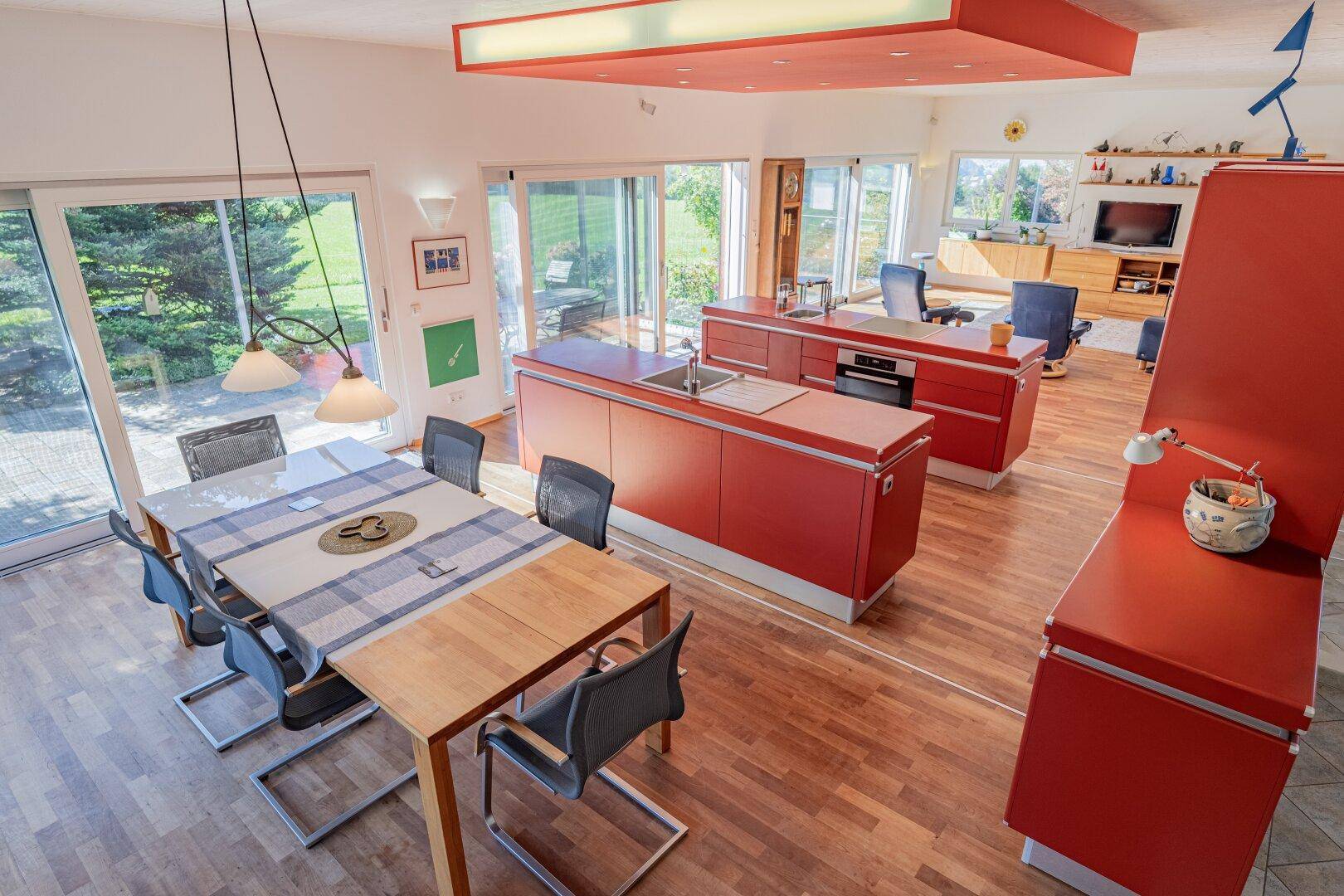 Offener Wohn-Essbereich mit moderner roter Küche, Holzboden und viel Tageslicht durch große Fenster.