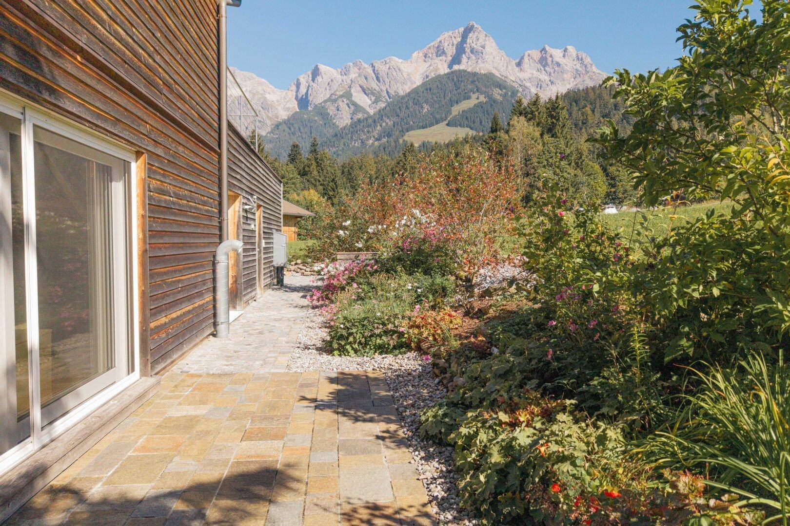 Gepflegter Gartenweg entlang des Holzhauses mit Blumenbeeten und Bergblick im Hintergrund.