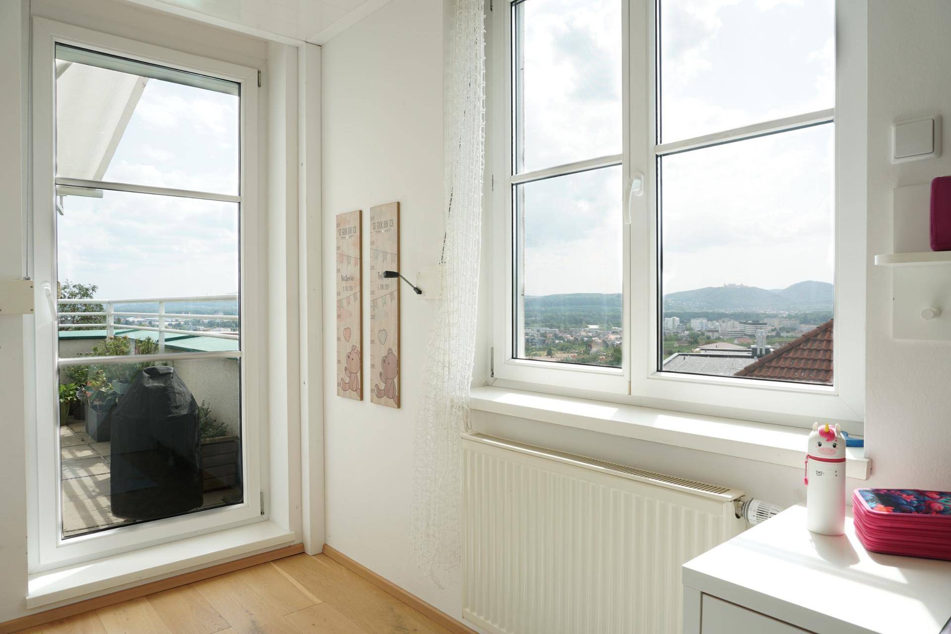 Heller Raum mit großen Fenstern, Blick auf die Stadt und Zugang zu einem Außenbereich.