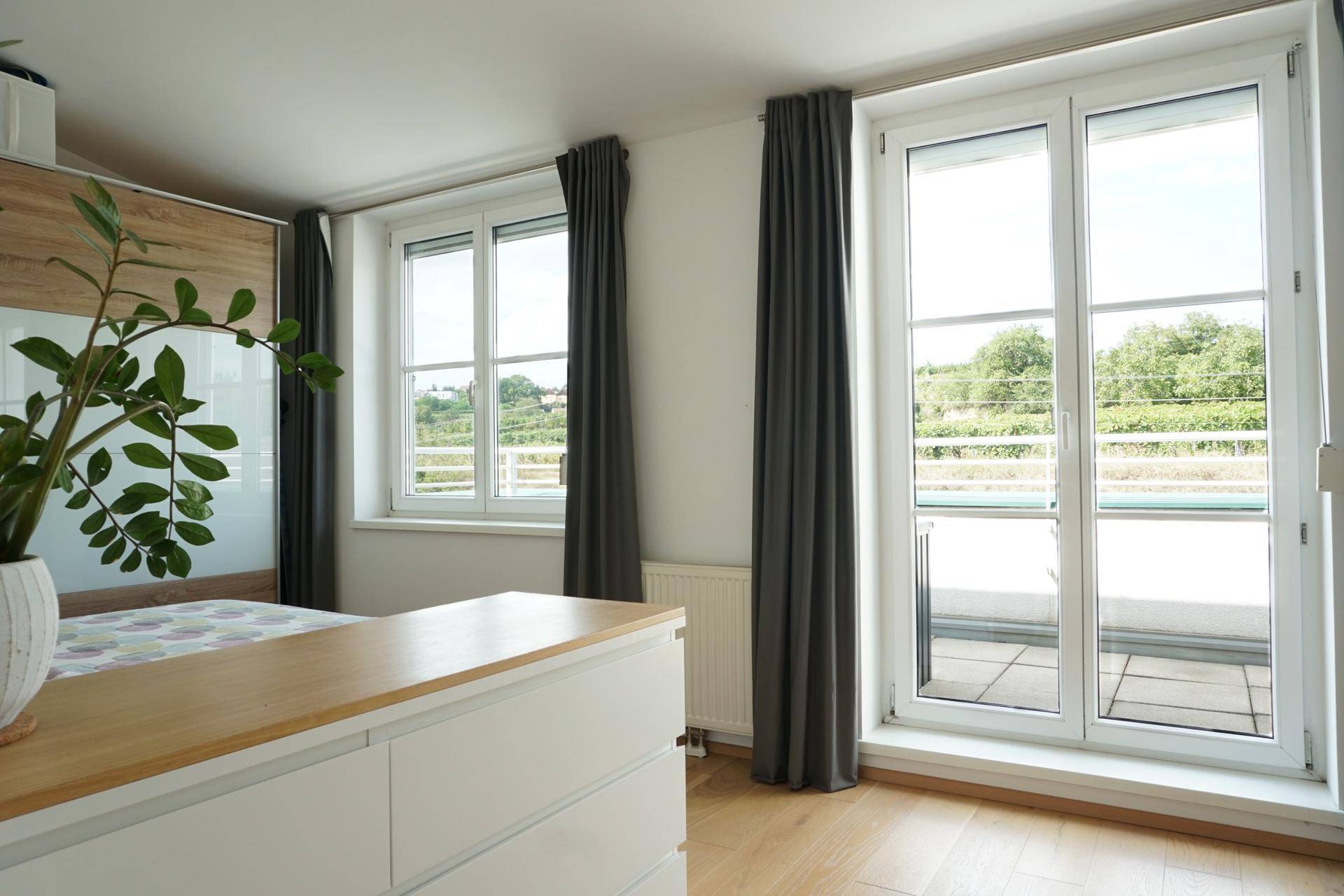 Helles Schlafzimmer mit Holzboden, Kommode und großen Fenstern mit Blick ins Grüne.