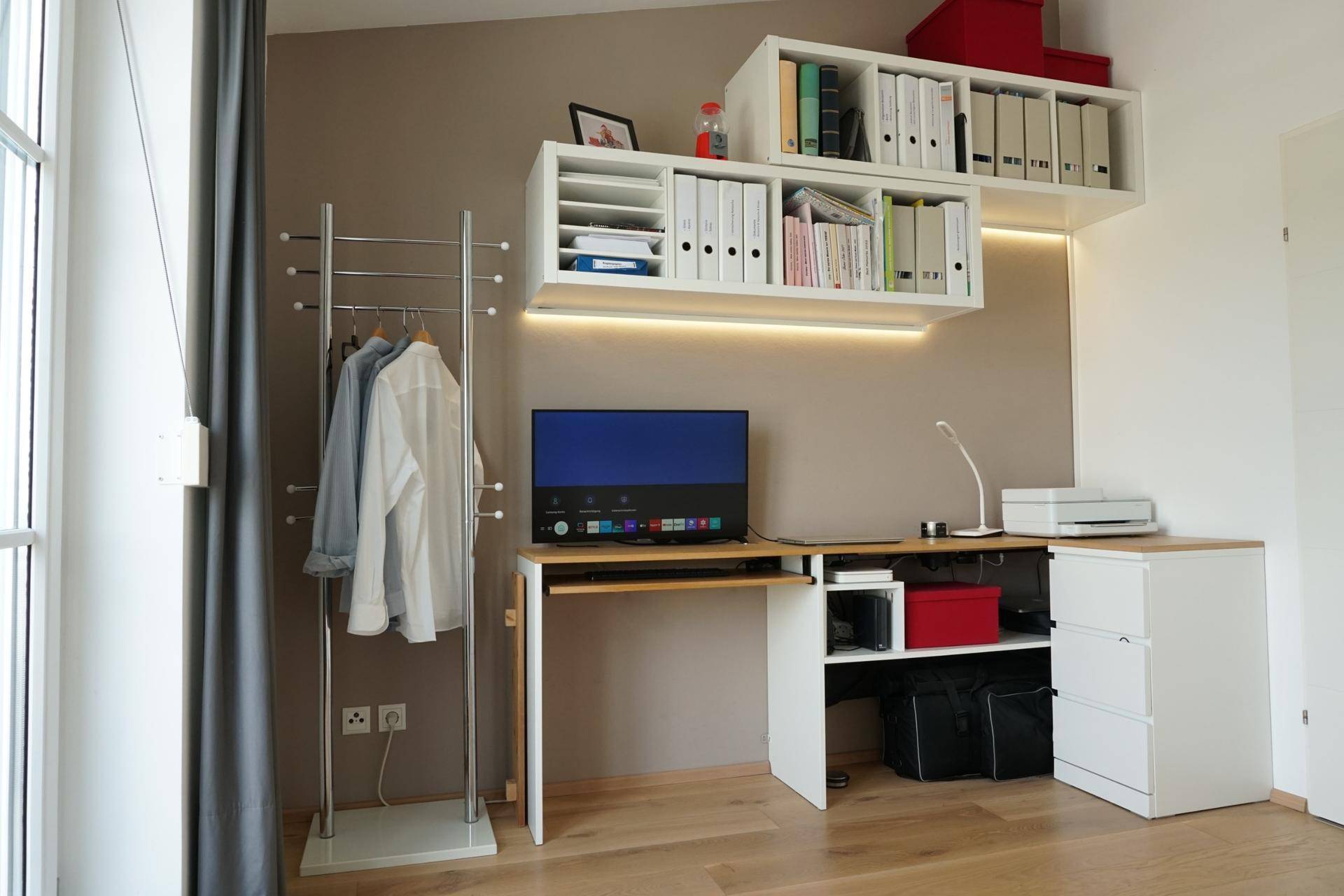 Funktionaler Arbeitsbereich mit Schreibtisch, Regalen und Kleiderständer, gut beleuchtet.
