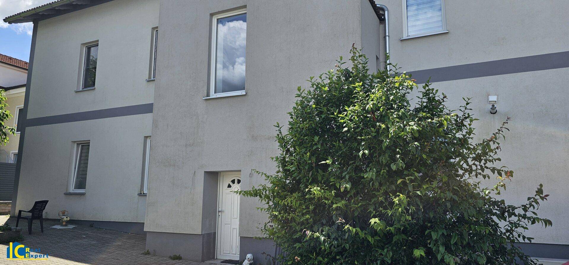 Seitenansicht des Gebäudes mit Eingangstür und üppigem Grün vor der Fassade unter blauem Himmel.