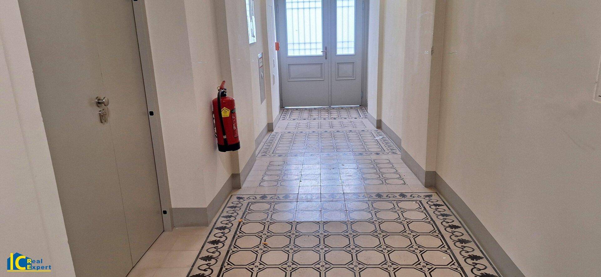 Heller Flur mit gemustertem Fliesenboden, Feuerlöscher und Eingangstür am Ende.