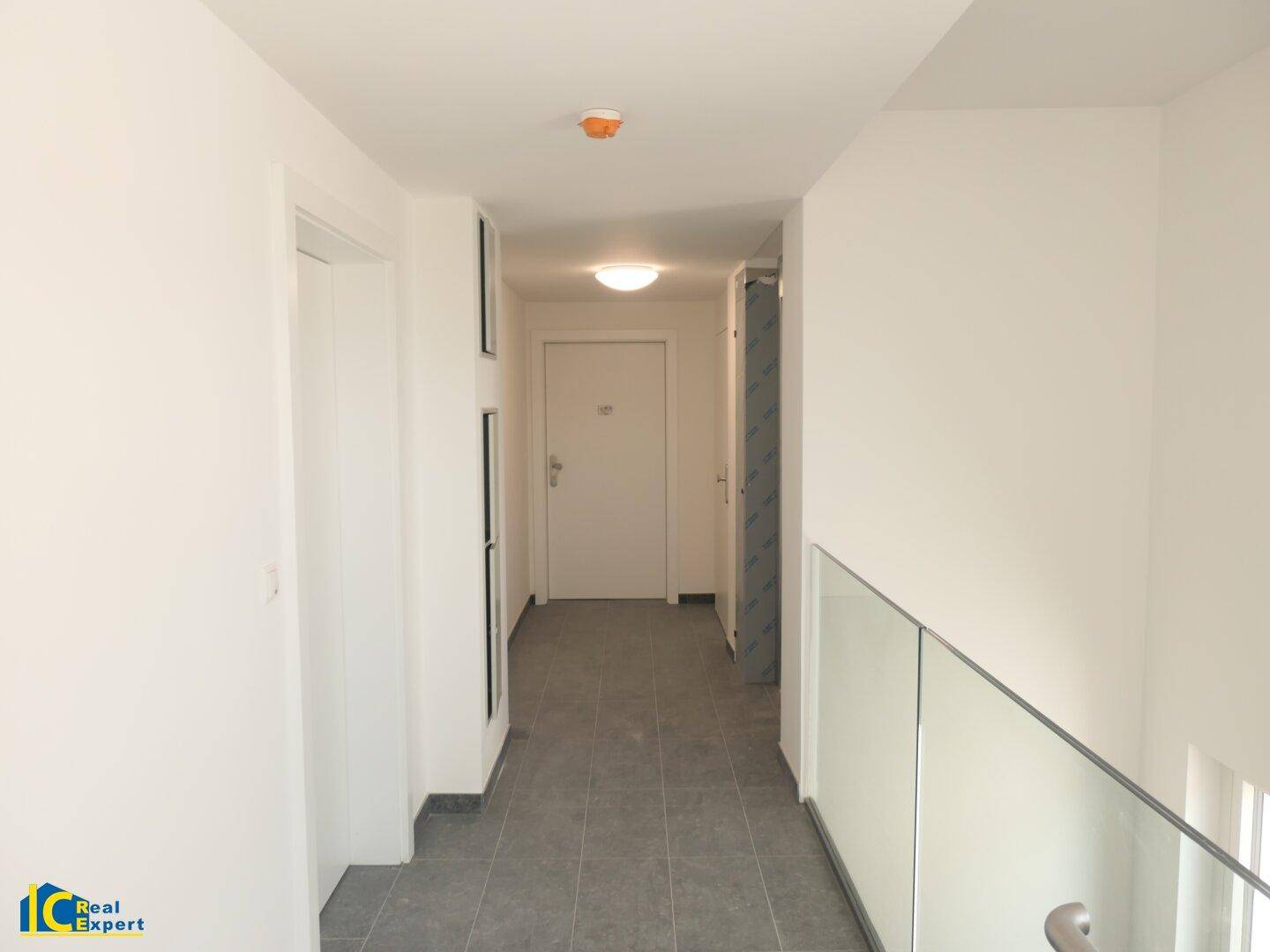 Moderner, heller Flur mit grauen Fliesen und Glasgeländer zu einem Treppenabgang.