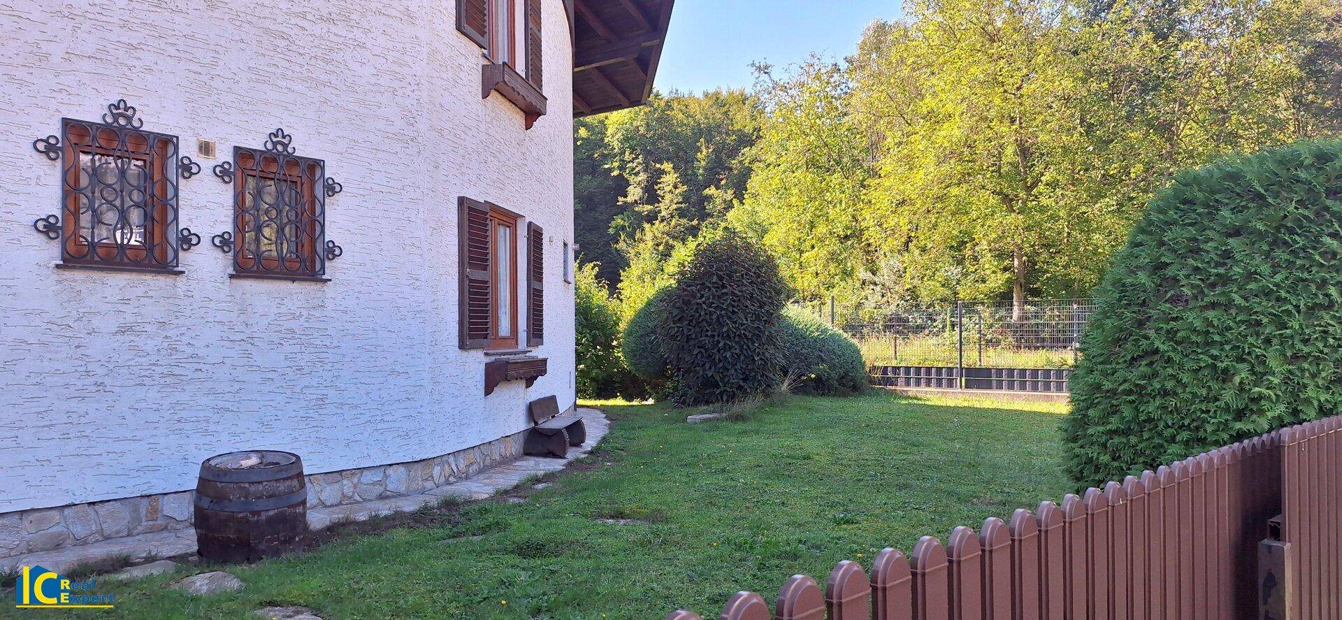 Seitlicher Gartenbereich des Hauses mit grüner Hecke und traditionellen Fenstergittern in ländlicher Umgebung.