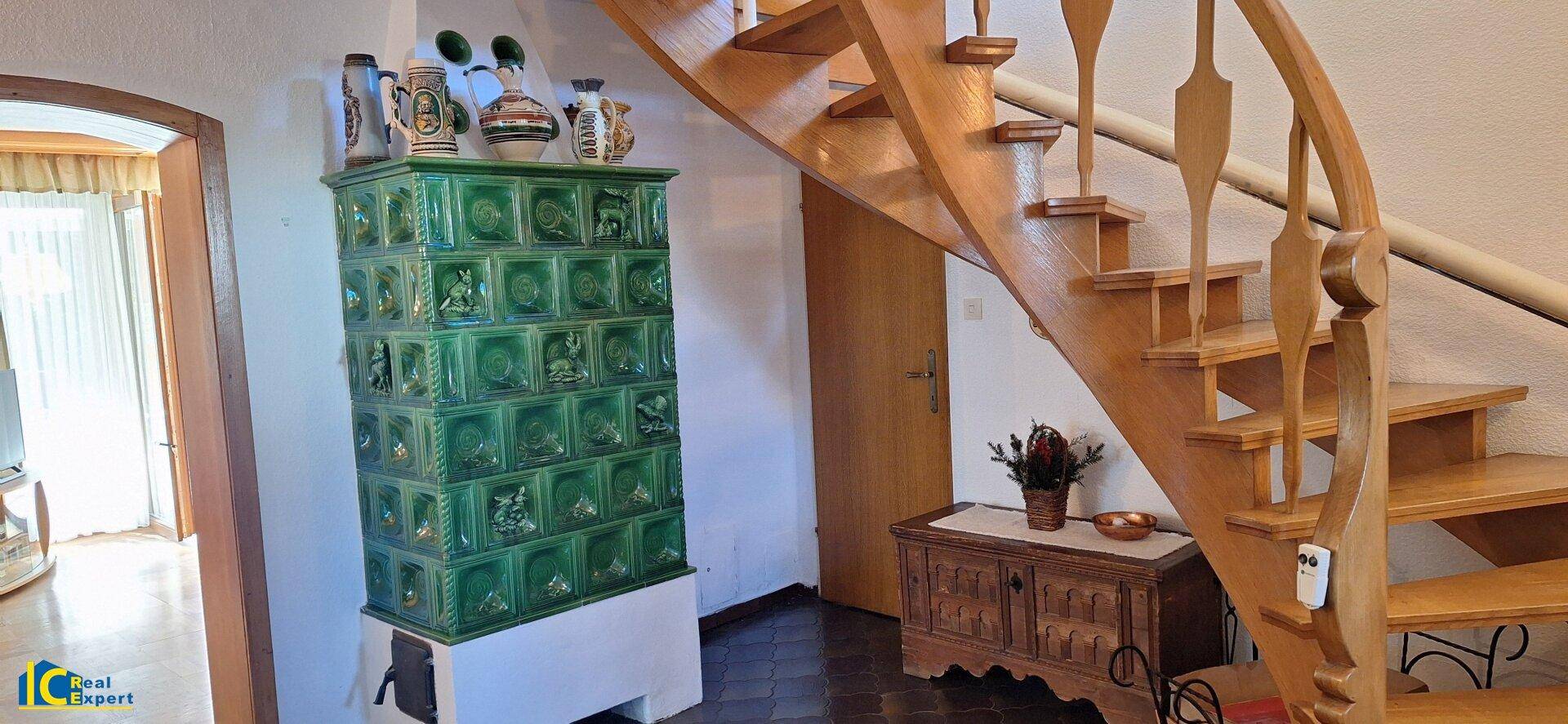 Geräumiger Flur mit Holztreppe und einem traditionellen grünen Kachelofen als Blickfang.