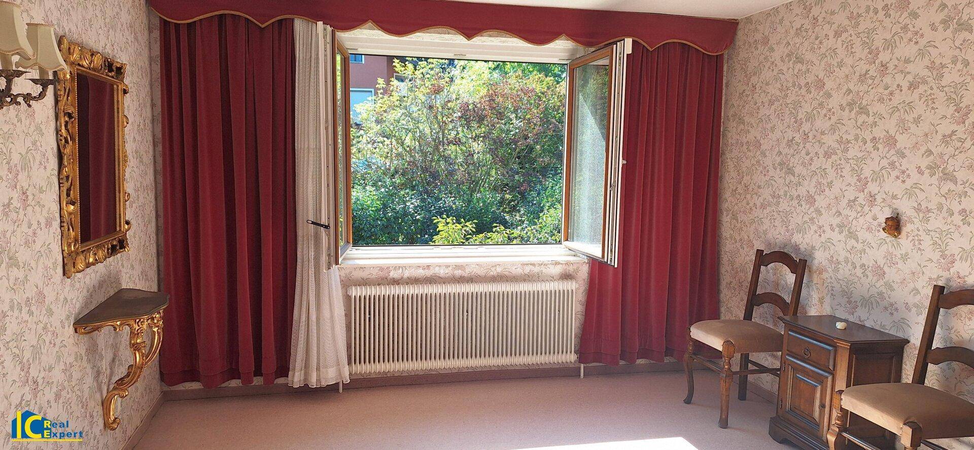 Schlafzimmer mit großem Fenster, rotem Vorhang und traditioneller Tapete, bietet Blick ins Grüne.