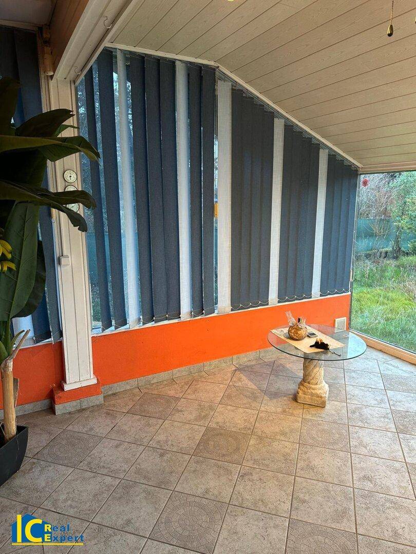 Heller Wintergarten mit Fliesenboden, orangefarbenen Wänden und vertikalen Jalousien für Privatsphäre.