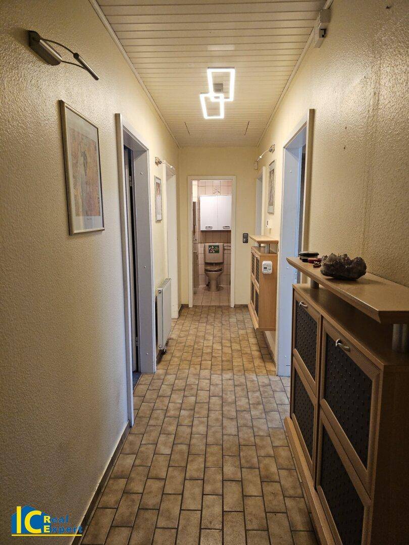 Langer Flur mit Fliesenboden, Holzmöbeln und moderner Deckenbeleuchtung, die den Raum erhellt.