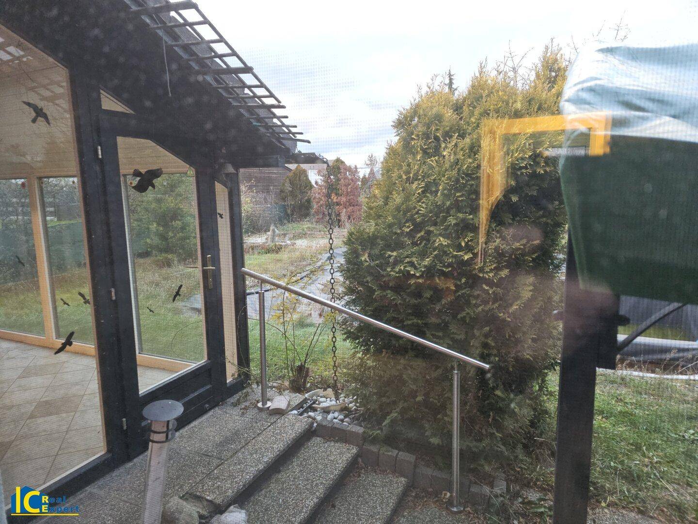 Verglaster Wintergarten mit Fliesenboden und Blick in den naturbelassenen Gartenbereich.