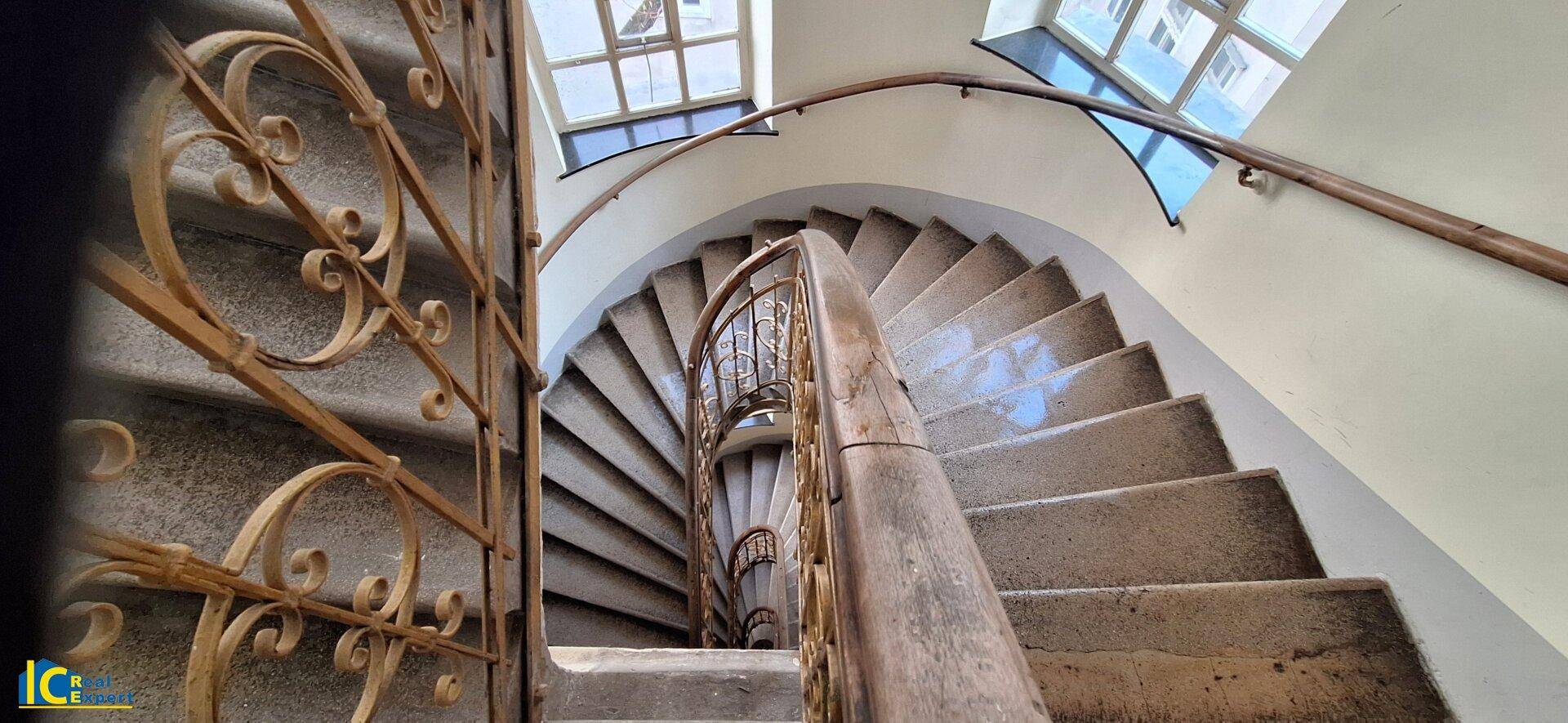 Elegantes Treppenhaus mit geschwungener Steintreppe, kunstvollem Metallgeländer und Fenstern für Tageslicht.