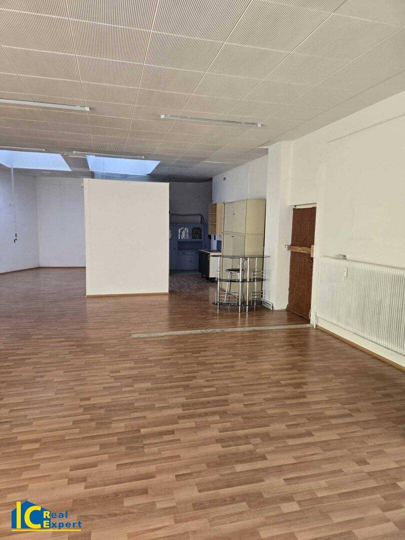 Großer, leerer Gewerberaum mit Laminatboden und Blick auf einen hinteren Bereich mit kleiner Küche.