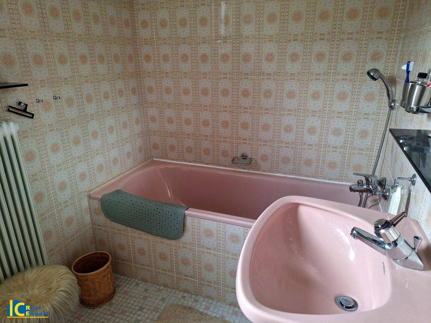 Badezimmer im 80er-Jahre-Stil mit rosa Badewanne, passendem Waschbecken und gemusterten Fliesen.