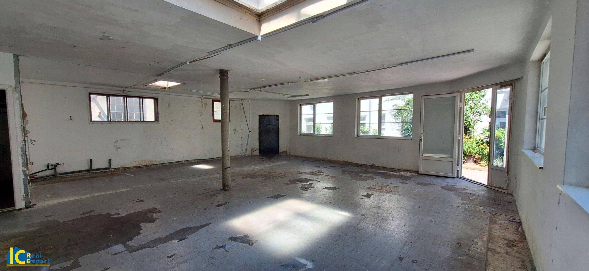 Geräumiger, leerer Raum mit Betonboden, vielen Fenstern und einer Glastür, die nach draußen führt.