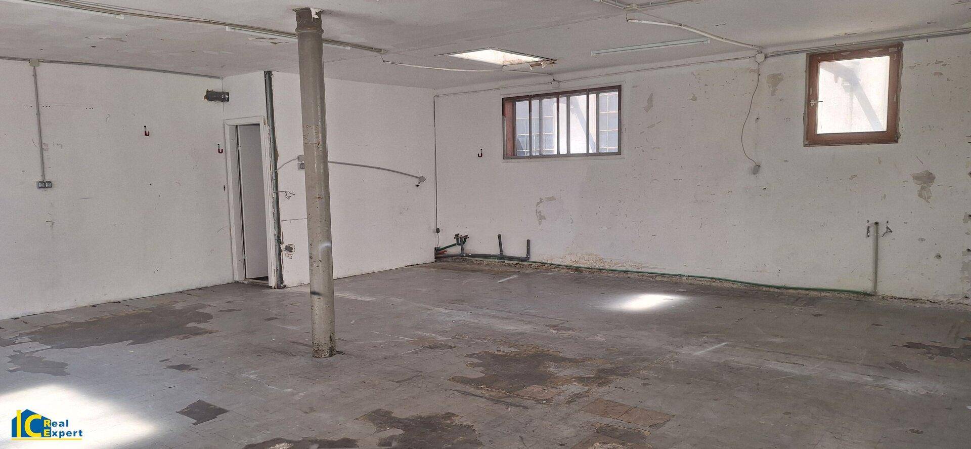 Blick in den hinteren Bereich des großen, leeren Raumes mit Betonboden und kleineren Fenstern.