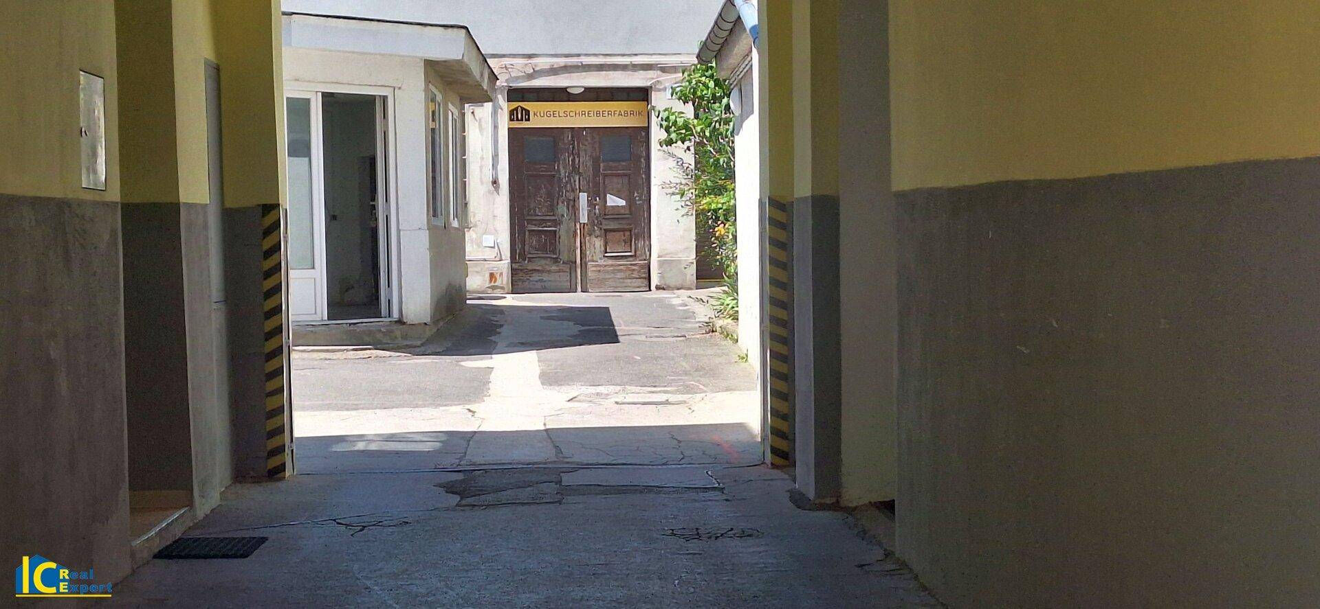 Blick durch eine gepflasterte Gasse oder einen Innenhof mit Gebäuden und einer großen Holztür am Ende.