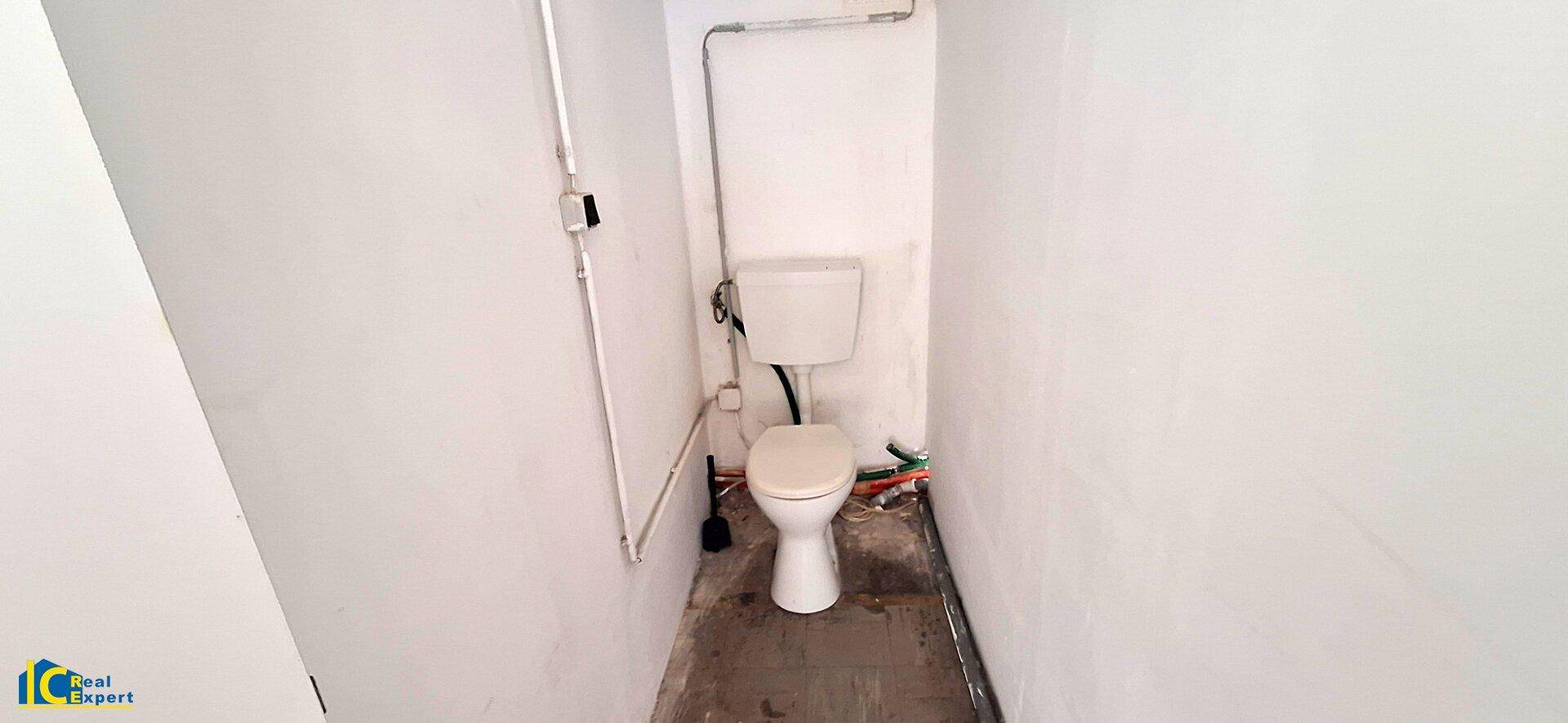 Kleiner Toilettenraum mit einfacher Toilette und sichtbaren Rohrleitungen, in schlichtem Zustand.