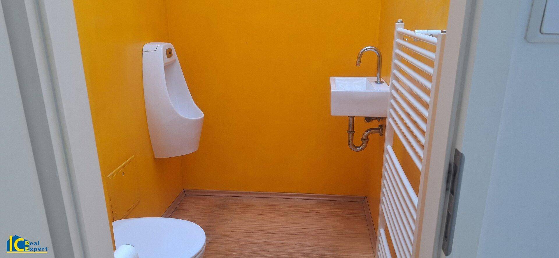 Kleiner Toilettenraum mit leuchtend gelben Wänden, Urinal, kleinem Waschbecken und Handtuchheizkörper.
