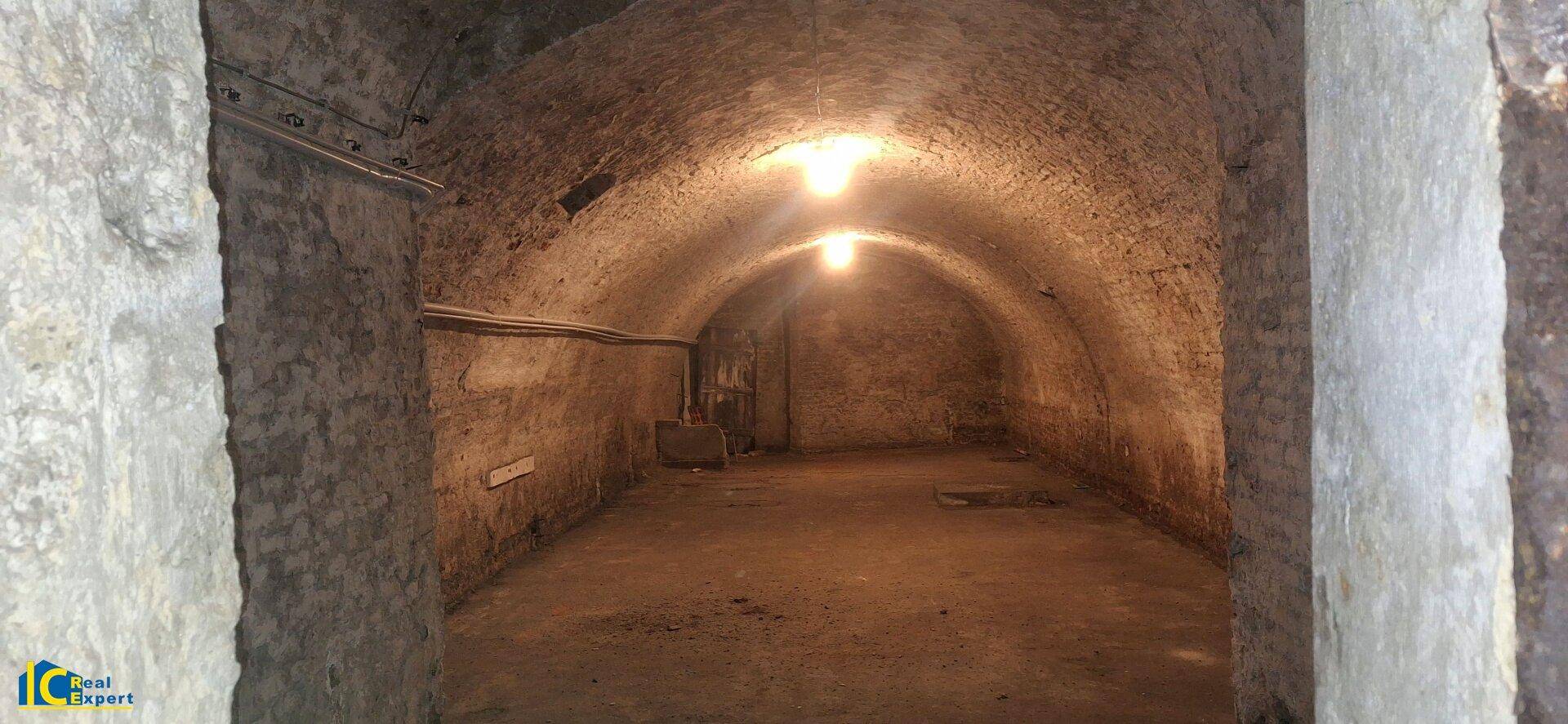 Alter, gewölbter Kellerraum mit Steinmauern und Betonboden, beleuchtet von zwei Glühbirnen.