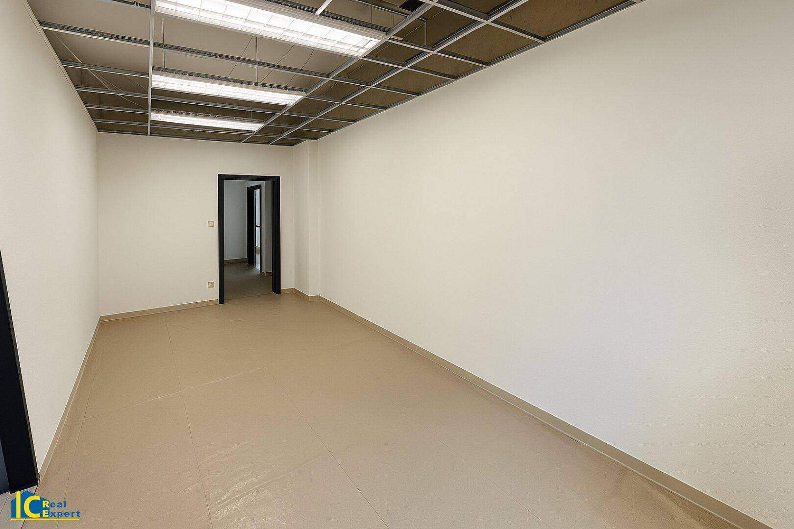 Leerer Büroraum mit beigem Vinylboden, weißen Wänden und sichtbarer Deckenkonstruktion.