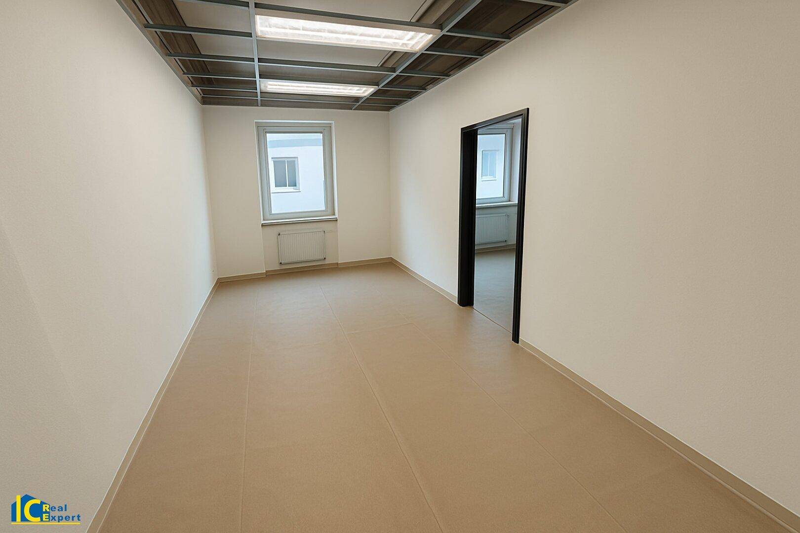 Heller Büroraum mit beigem Vinylboden, weißen Wänden und einem Fenster mit Heizkörper.
