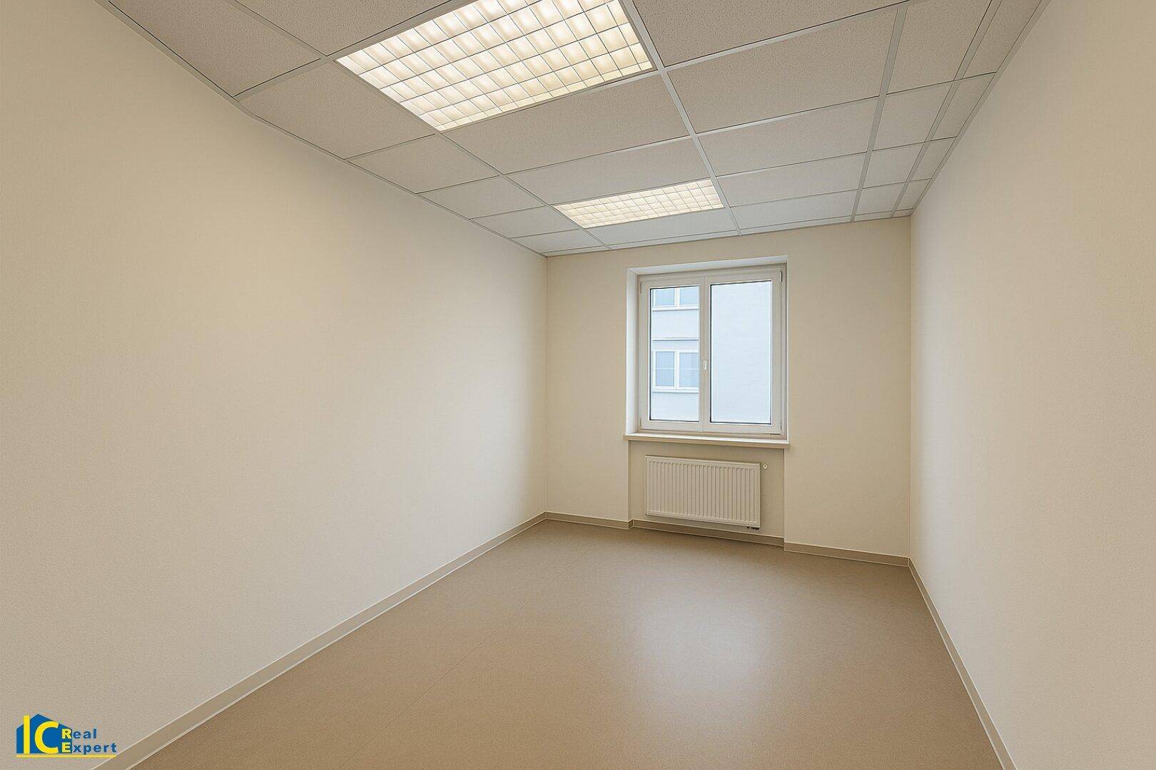 Leerer Büroraum mit beigen Wänden, abgehängter Decke und einem Fenster mit Heizkörper.