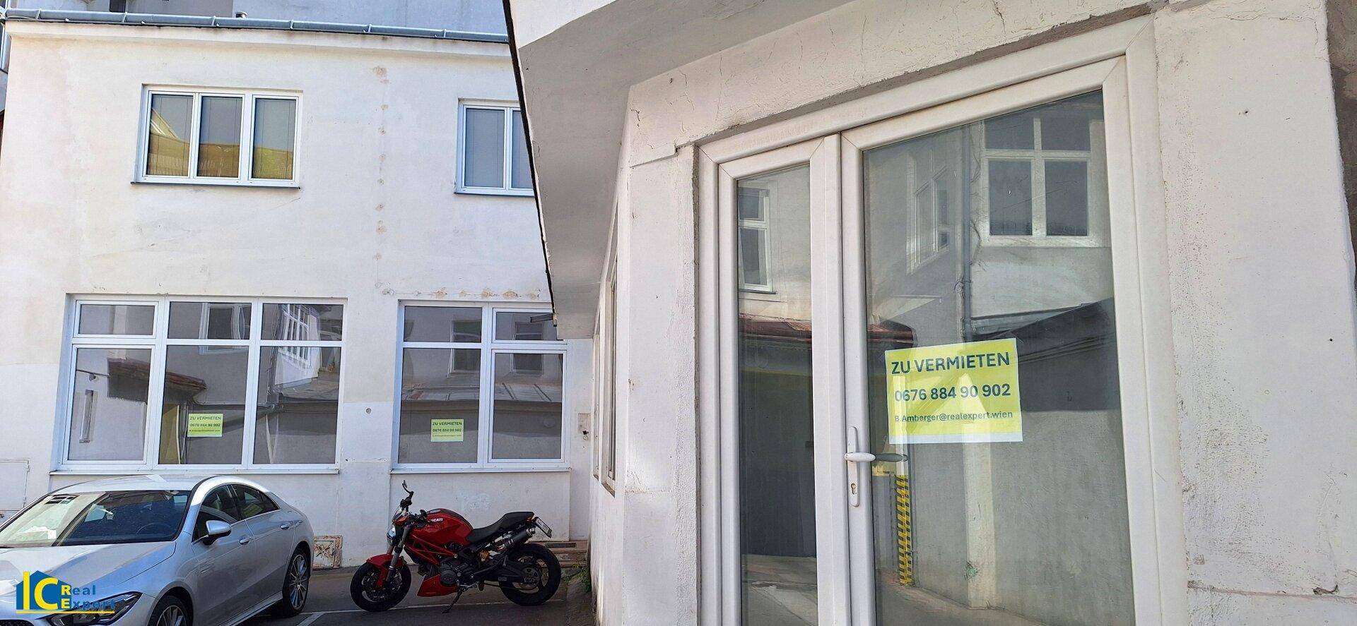 Eingangsbereich des kleinen Gebäudes mit einem "Zu vermieten"-Schild an der Glastür und geparkten Fahrzeugen.