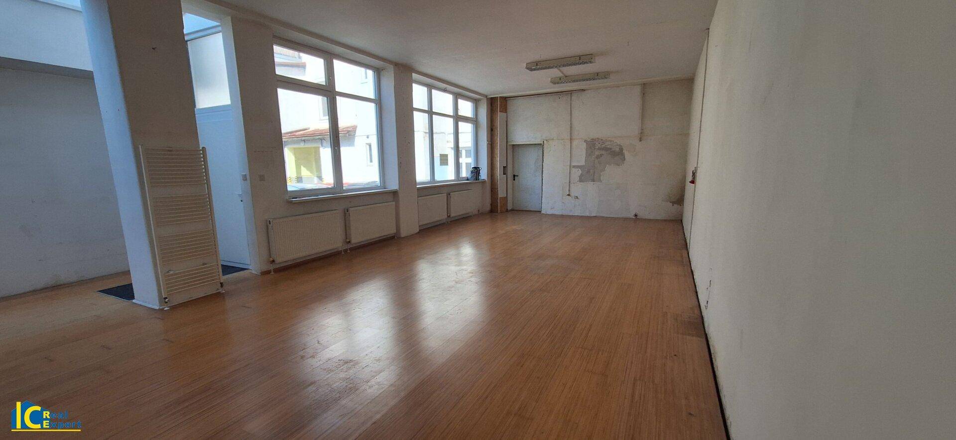 Langer Raum mit Holzplankenboden, einer Reihe großer Fenster und Heizkörpern, ideal für Büronutzung.