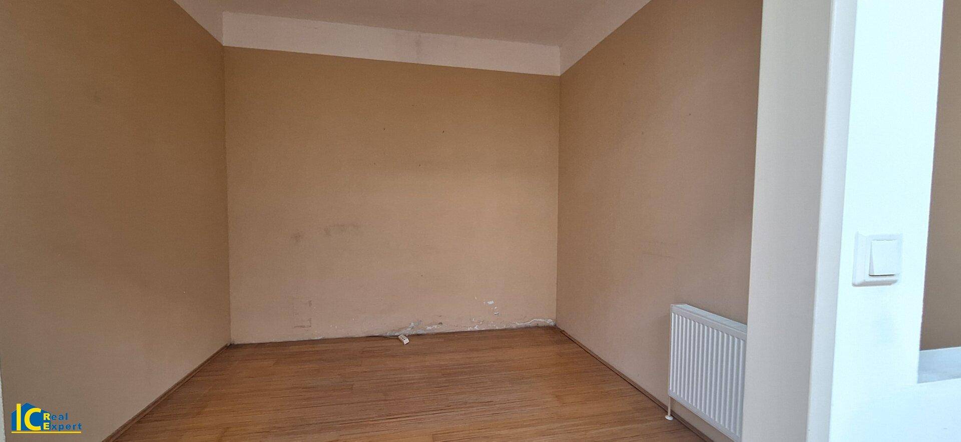 Kleiner, leerer Raum mit beigen Wänden, Holzplankenboden und einem Heizkörper, bereit zur Nutzung.