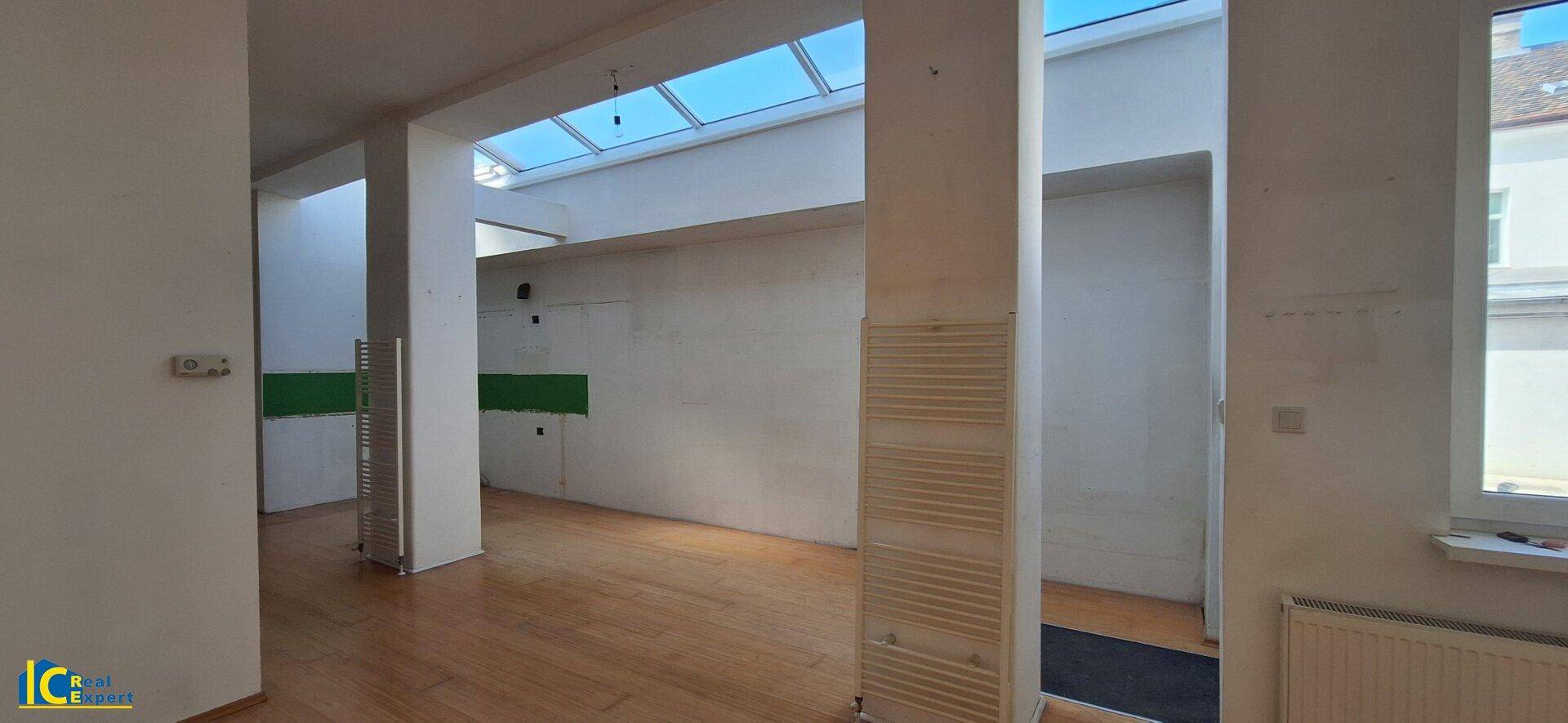 Heller Raum mit Holzplankenboden, markanten Säulen und einem großen Oberlicht, das den Raum erhellt.