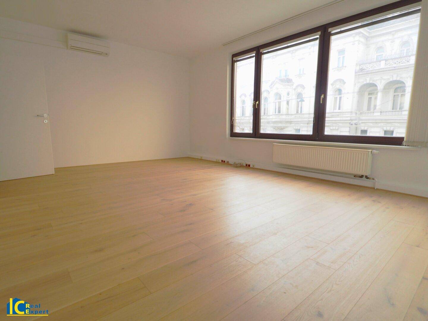Heller, leerer Raum mit großen Fenstern, hellem Holzfußboden und Blick auf ein gegenüberliegendes Gebäude.