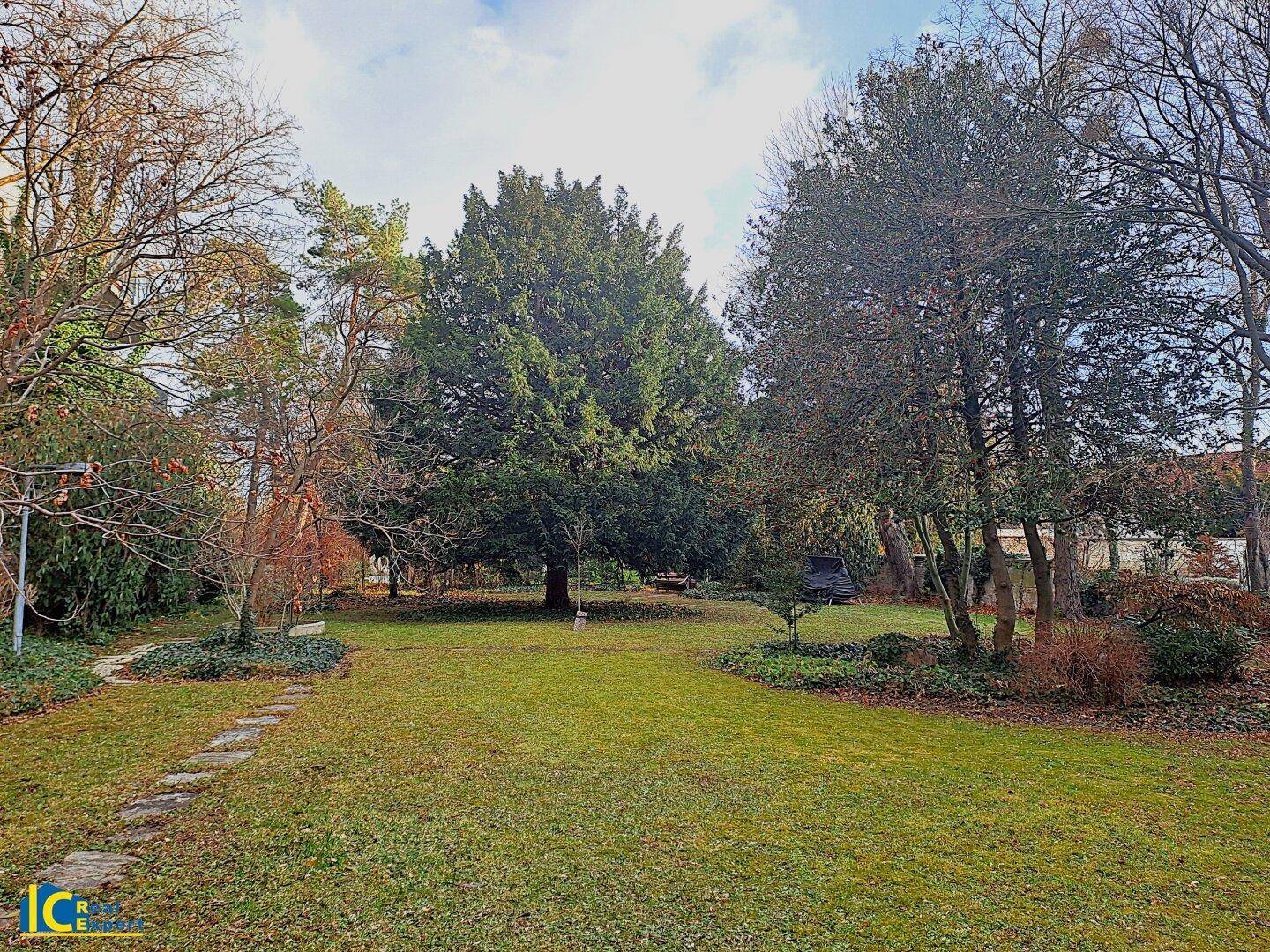Großer, gepflegter Garten mit altem Baumbestand und weitläufiger Rasenfläche unter bewölktem Himmel.