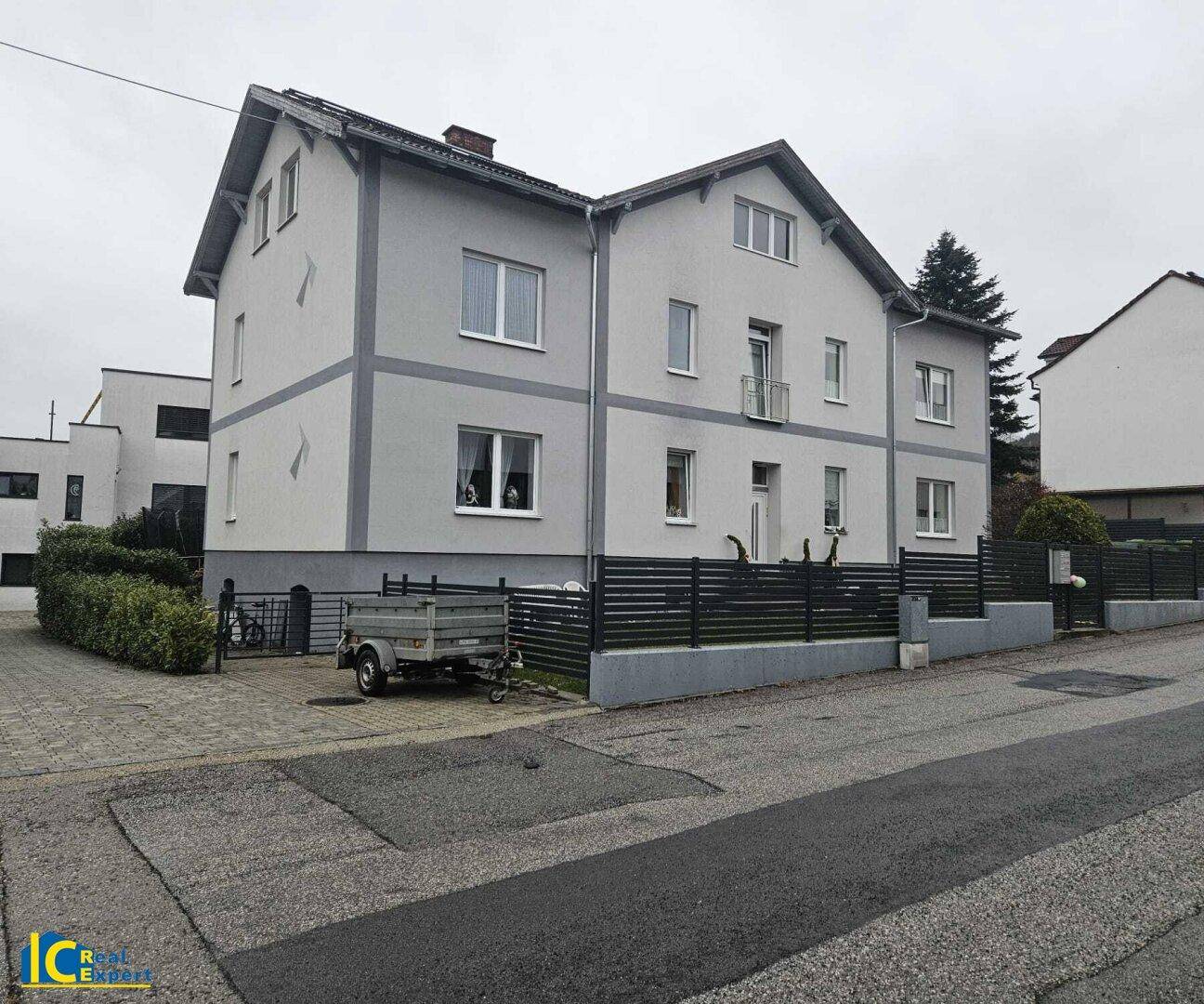 Gesamtansicht des grauen Wohnhauses mit Vorgarten, Anhänger und der angrenzenden Straße.