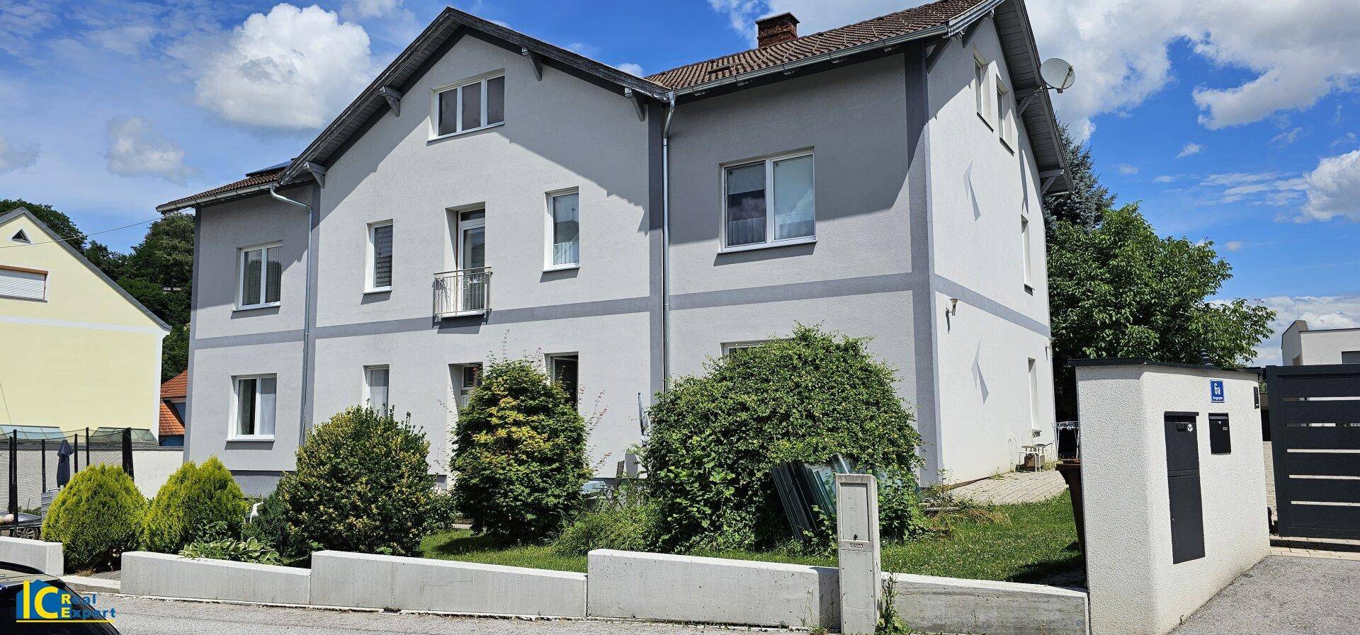 Ansicht des grauen Wohnhauses mit gepflegtem Vorgarten und blauem Himmel an einem sonnigen Tag.