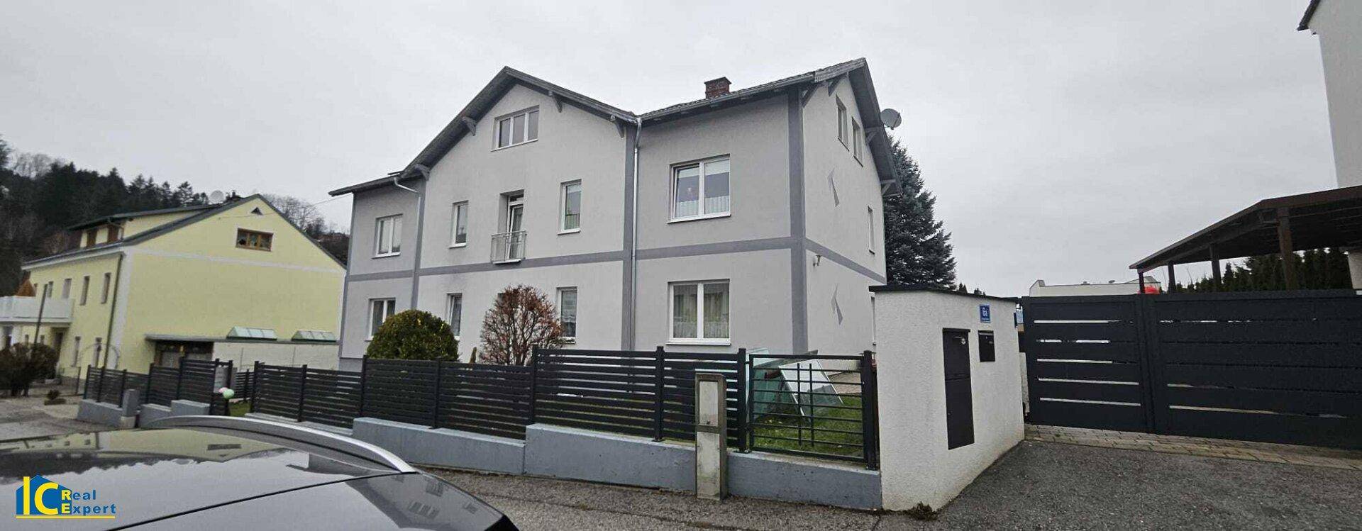 Außenansicht des grauen Wohnhauses mit Zaun und Einfahrt an einem bewölkten Tag.