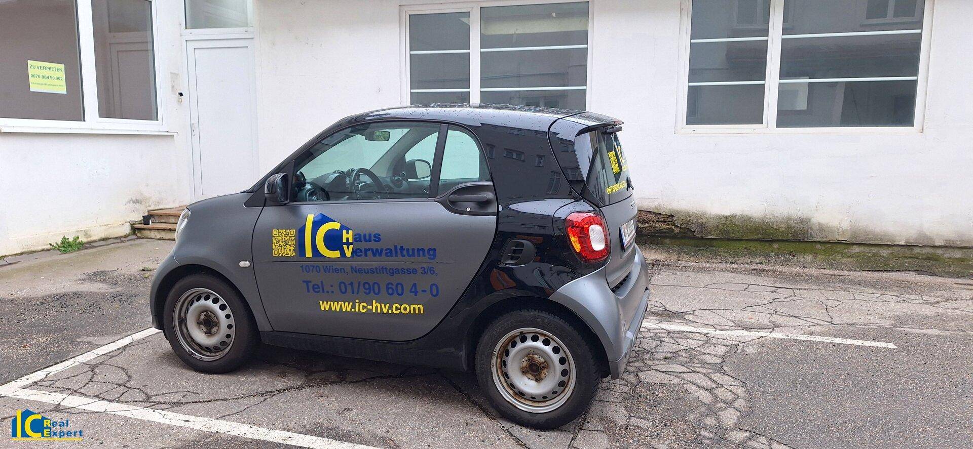 Ein kleines Auto mit Werbung parkt auf einem gepflasterten Bereich neben einem Gebäude.