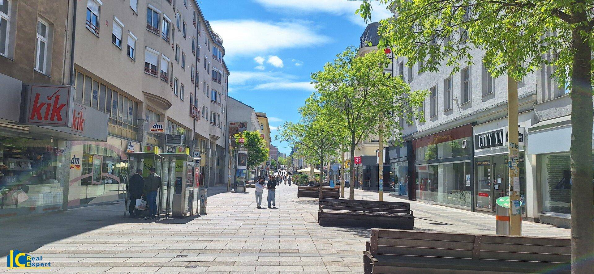 Belebte Einkaufsstraße mit Geschäften, Bänken und Fußgängern unter blauem Himmel.