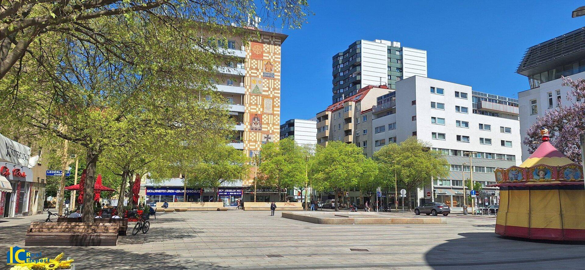 Belebtes Stadtzentrum mit modernen Gebäuden, Bäumen und einem Karussell.