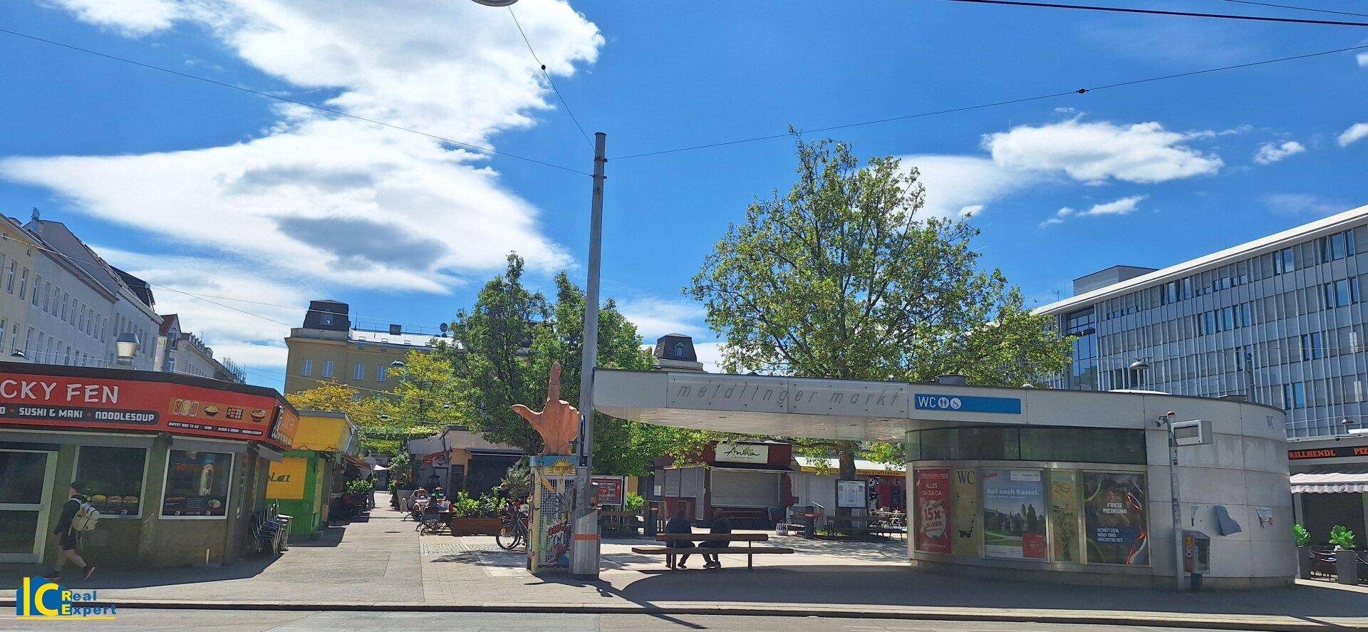 Öffentlicher Bereich mit Geschäften, Bäumen und einem U-Bahn-Eingang.