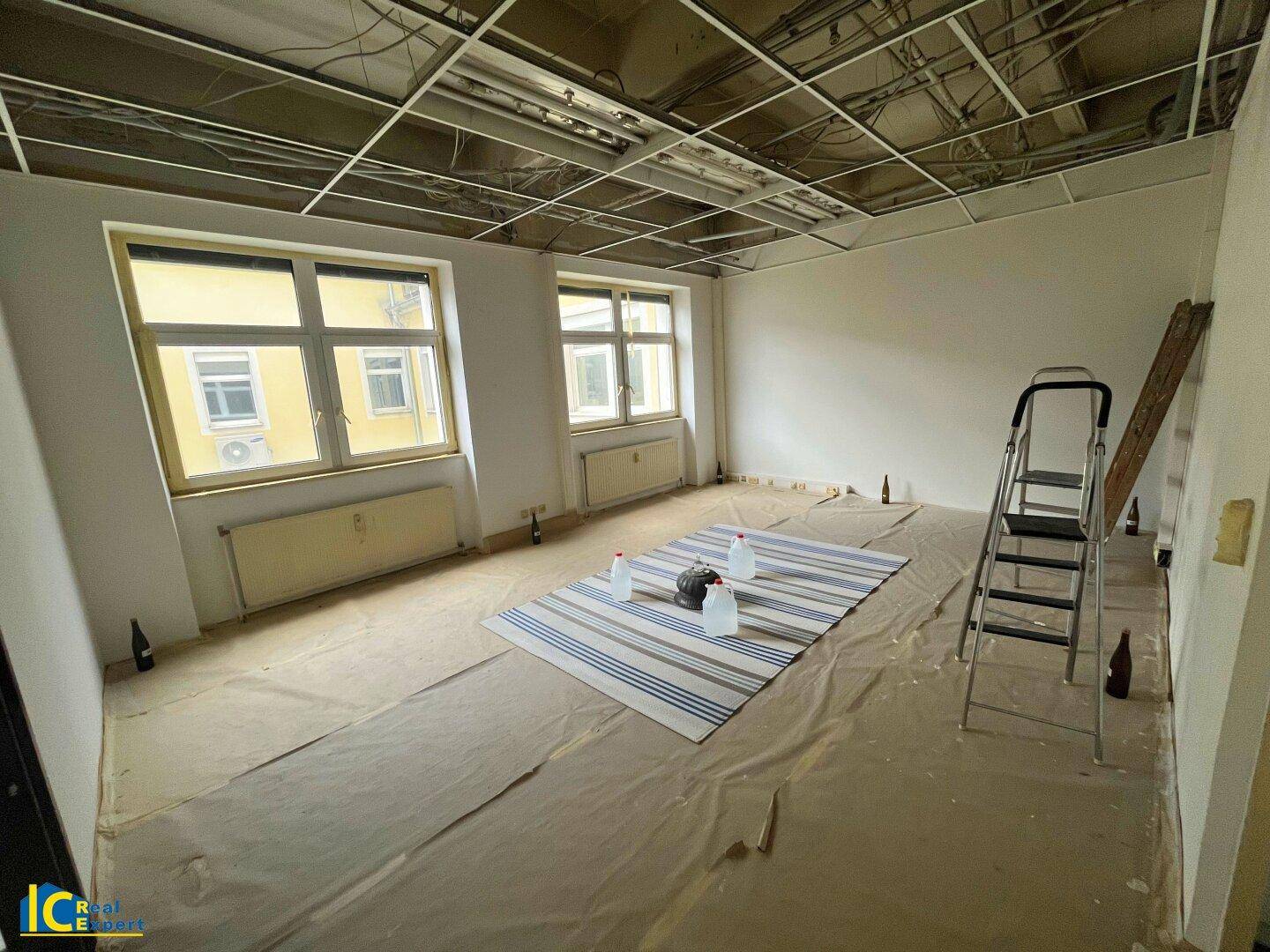 Unfertiger Innenraum mit abgehängter Decke, zwei großen Fenstern und Schutzfolie auf dem Boden während der Renovierung.