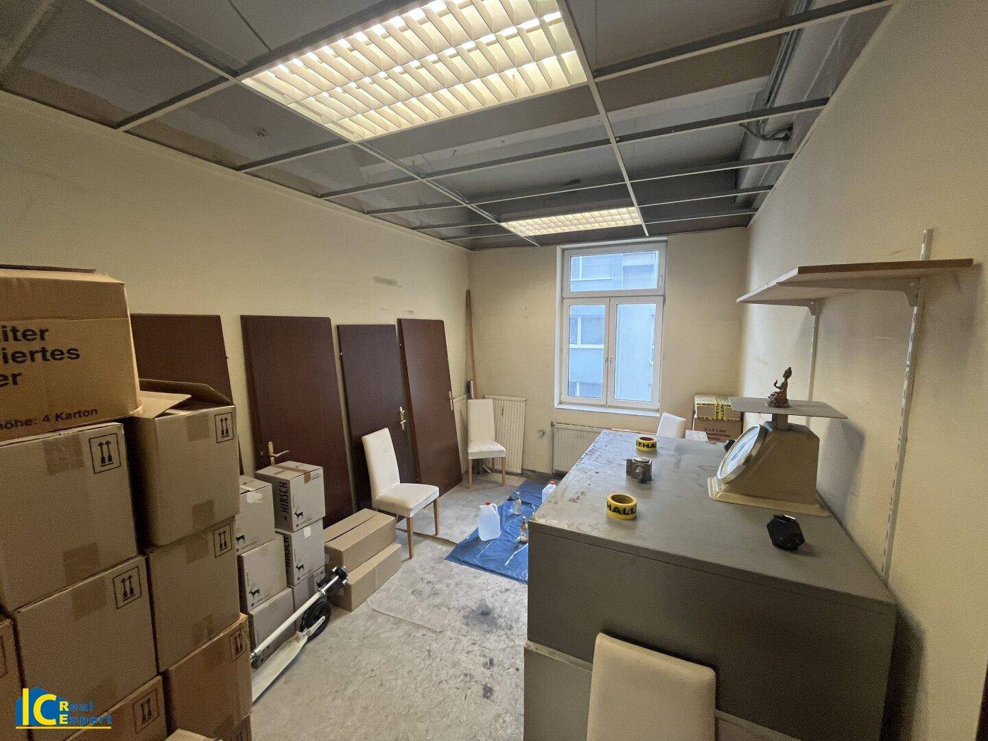 Renovierungsbedürftiger Raum mit Kartons, alten Möbeln, einer Waage und einem Fenster, das Tageslicht hereinlässt.